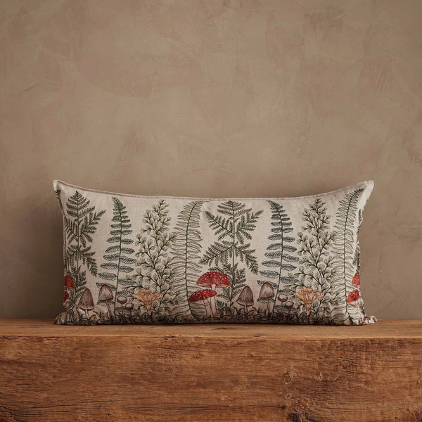 The Coral & Tusk Mushrooms and Ferns Lumbar Pillow is a rectangular embroidered linen accent featuring botanical mushrooms and ferns in earthy tones, displayed on a rustic wooden surface against a beige textured wall.