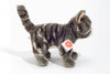 The Teddy Hermann Standing Tabby Cat Stuffed Animal, with its gray and white striped fur, stands prominently on a white surface. This high-quality plush toy features a tag on its ear displaying the distinctive red and gold label from the Hermann Teddy Collection. Positioned in profile, it faces right with its tail raised in the air.