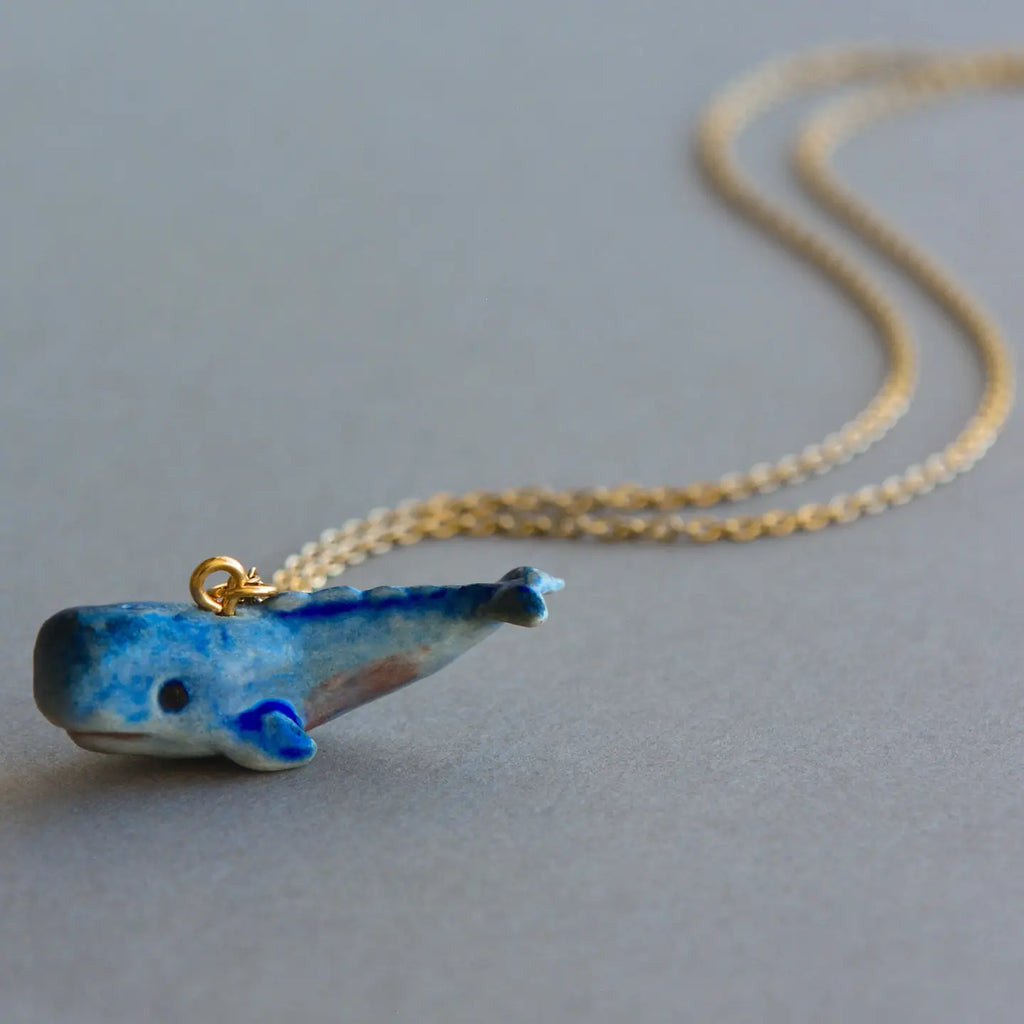 A Blue Whale Necklace featuring a small, ceramic pendant hand painted in detail. The whale, captured with smiling expression and aquamarine tones, is set against an elegantly laid out necklace chain on a neutral gray surface.