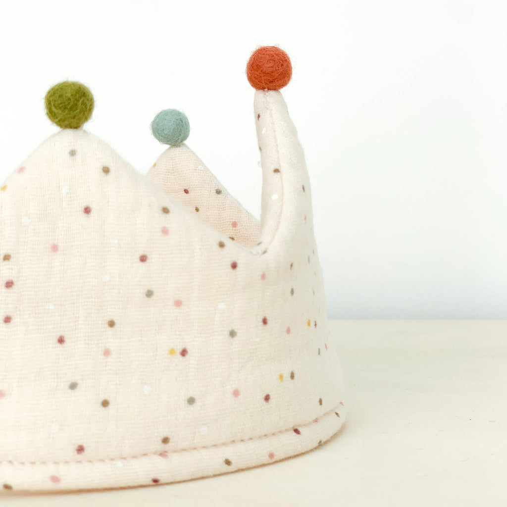 A Confetti Reversible Birthday Crown with pastel polka dots and three points, each topped with a colorful felt ball (green, teal, and orange). Crafted from organic cotton, the reversible crown rests on a light wooden surface against a plain white background.