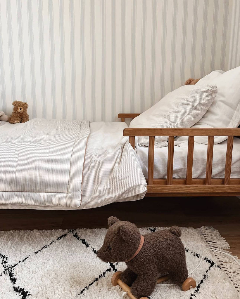 A cozy child's bedroom features a wooden bed with striped bedding, a small teddy bear on the bed, and the Pull-Along Raoul toy placed on a white rug with black lines. The wall has light vertical stripes.