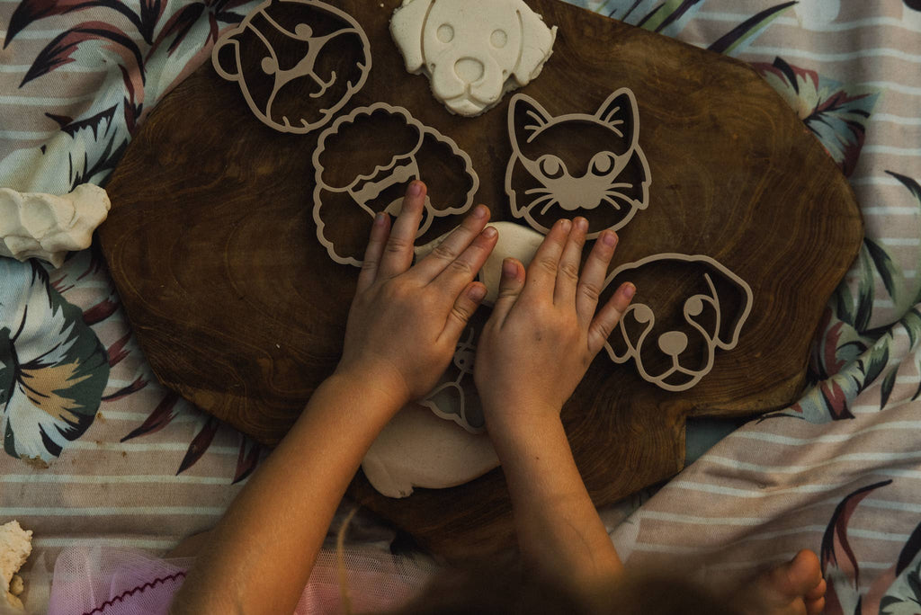 A child presses their hands onto the Pet Friends Faces Cutter Set – Eco Playdough Tool, shaping dough on a wooden board and transforming sensory play into a delightful activity. The scene is arranged atop patterned fabric.