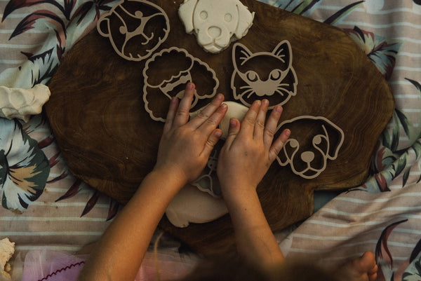 A child presses their hands onto the Pet Friends Faces Cutter Set – Eco Playdough Tool, shaping dough on a wooden board and transforming sensory play into a delightful activity. The scene is arranged atop patterned fabric.
