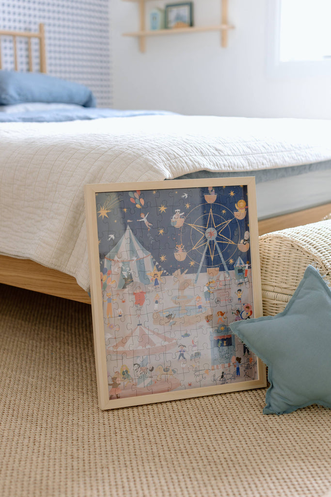 A Carnival of Wonders 100 Piece Puzzle, crafted from recycled materials for kids ages 5+, features a whimsical carnival scene in a cozy, sunlit bedroom. Nearby, a blue star-shaped pillow rests on the beige carpet.