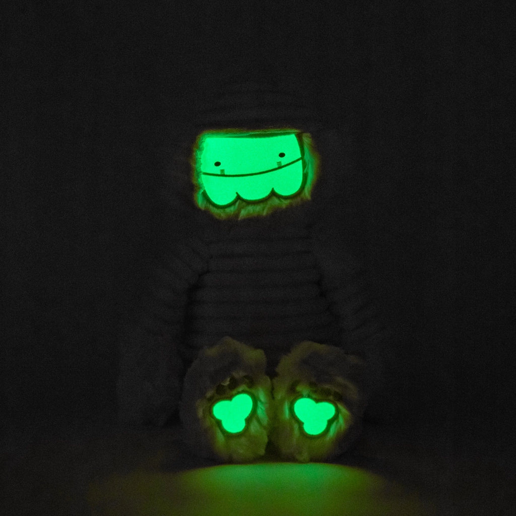 A glowing green figure in darkness, with a smiley face on its head and two clover-shaped eyes on its knees, resembling cartoonish features of a Slumberkins Halloween Gift Set - Mummy Kin + Halloween Fright Book.