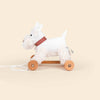 Pull-Along Lola is a white plush dog with a red-checkered collar, mounted on four wooden wheels with a pull string. Shown against a plain beige background, it encourages developmental play and supports gross motor skills.
