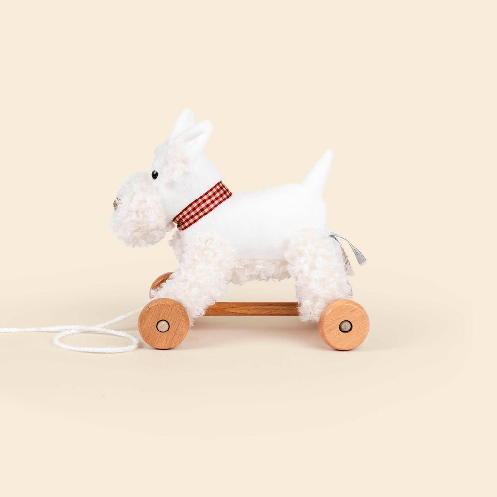 Pull-Along Lola is a white plush dog with a red-checkered collar, mounted on four wooden wheels with a pull string. Shown against a plain beige background, it encourages developmental play and supports gross motor skills.