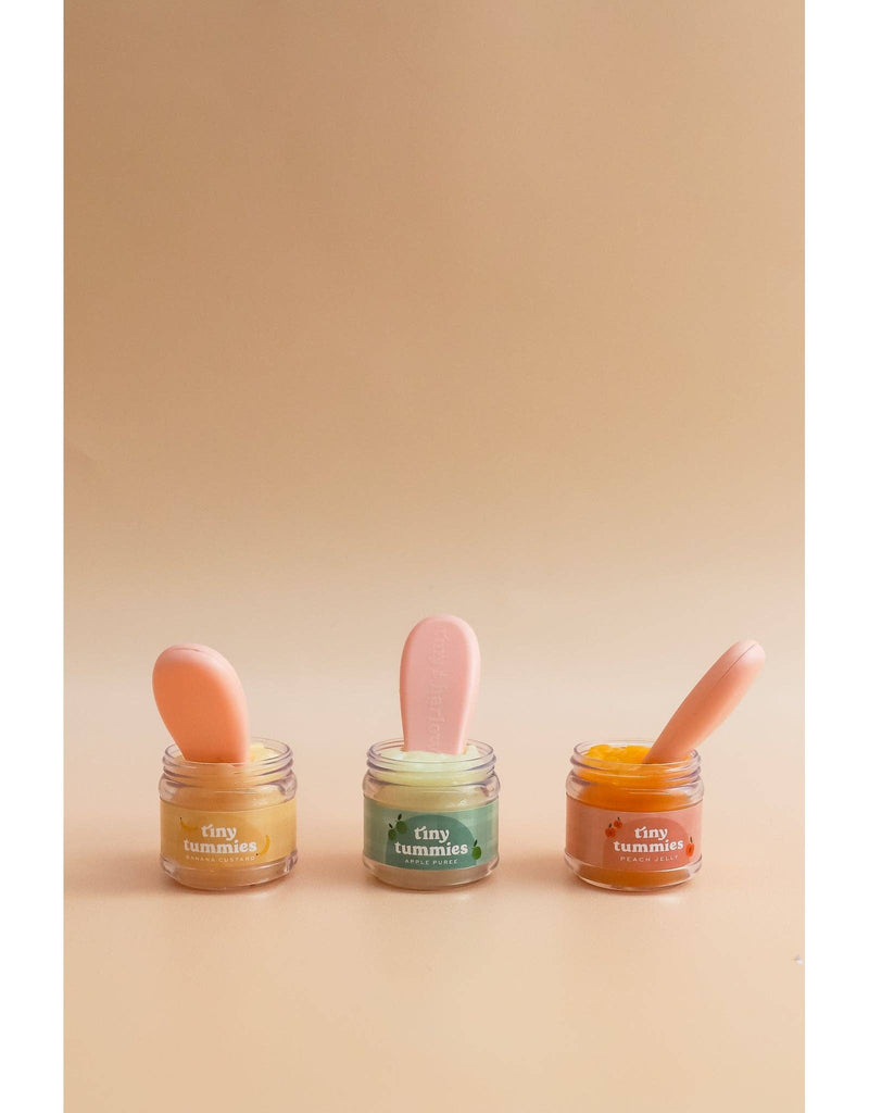 Three small jars labeled "Tiny Tummies" from Tiny Harlow contain peach jelly food in vibrant colors—yellow, green, and orange. Each jar includes a magical spoon sticking out against a warm beige background.