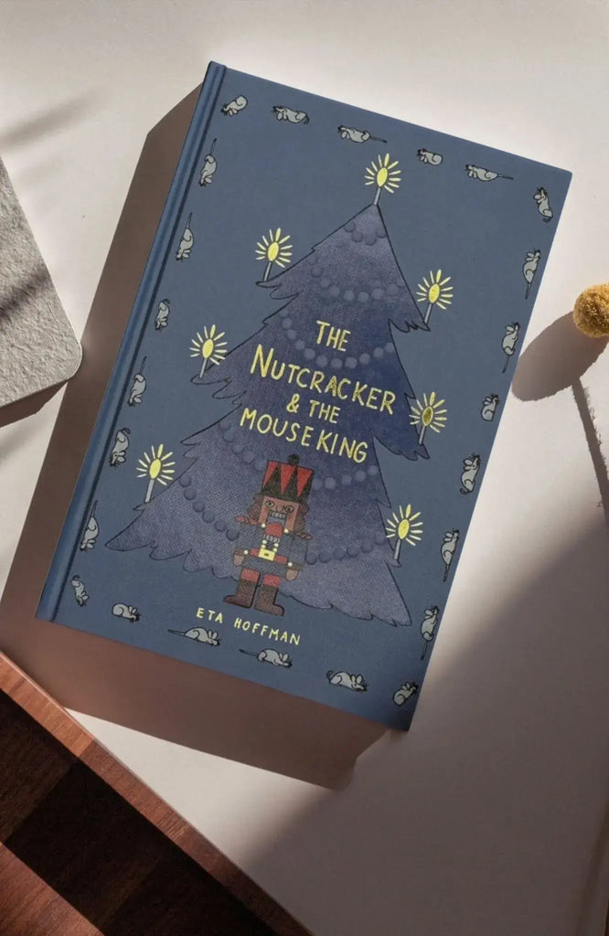 A hardcover copy of "The Nutcracker | Collector's Edition" lies on a table, featuring a blue cover adorned with a Christmas tree, nutcracker illustration, and small mice along the border.