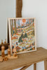 The Secrets of the Autumn Woods 300 Piece Puzzle, depicting a vibrant outdoor picnic scene with families enjoying nature, is displayed beside wooden toys, geometric shapes, and an eco-friendly reference poster.