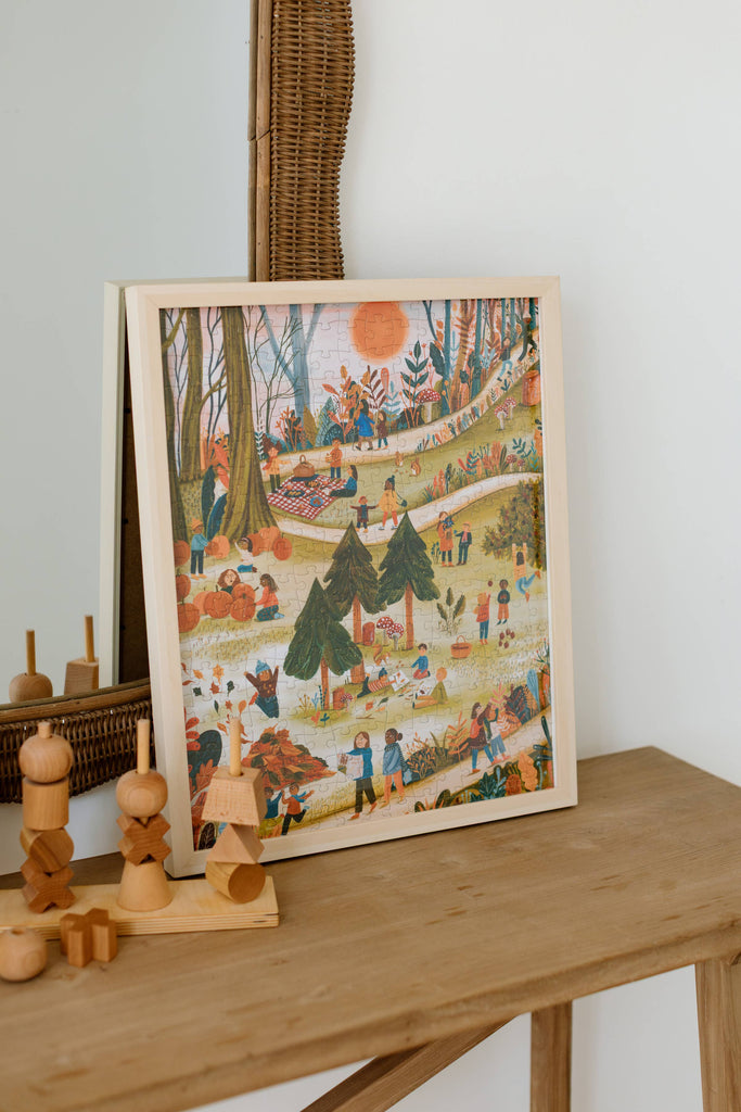 The Secrets of the Autumn Woods 300 Piece Puzzle, depicting a vibrant outdoor picnic scene with families enjoying nature, is displayed beside wooden toys, geometric shapes, and an eco-friendly reference poster.