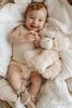 A smiling baby cuddles Snuggle Bunnies - Hilda The Highland Cow, the perfect plush companion and an adorable baby shower gift, while lying on a soft blanket and wearing a beige outfit with a matching headband.