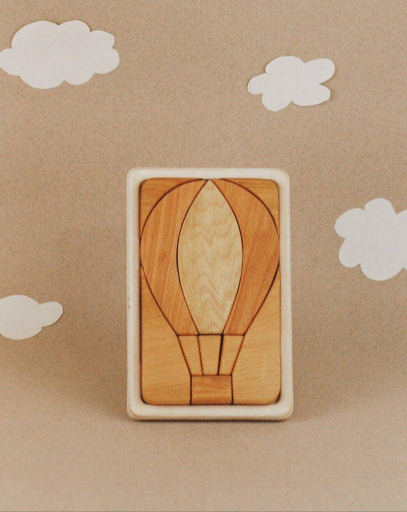 The Handmade Hot Air Balloon Puzzle is crafted from cherry wood and features a hot air balloon design against a beige background with white paper clouds, with puzzle pieces in varied shades showcasing intricate designs.