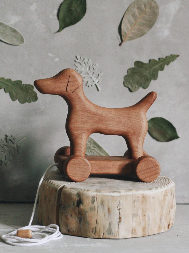 A Handmade Wooden Dog Pull Toy stands atop a tree stump. Handcrafted with simple carved features, the dog rests on four round wheels. A white pull string is attached to the front. The background includes various green leaves adhered to a gray wall.