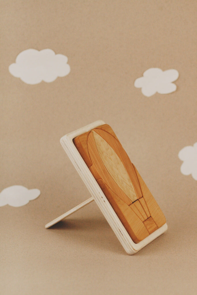 The Handmade Hot Air Balloon Puzzle, crafted from Cherry wood, sits elegantly on a detachable kickstand against a beige background, surrounded by whimsical white paper clouds.