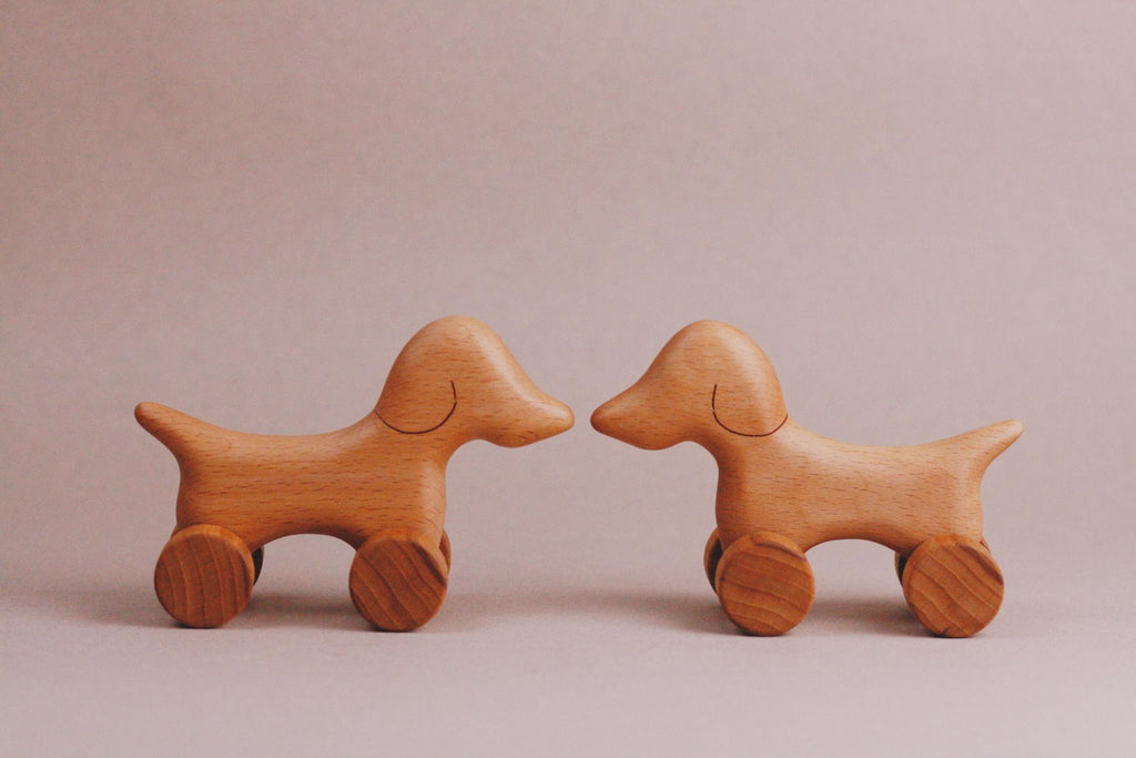 A pair of charming Handmade Wooden Mini Dog Push Toys, crafted from maple and beech wood, are positioned facing each other on a neutral background. These delightful figures feature wheels for legs, evoking playful creativity and timeless elegance.