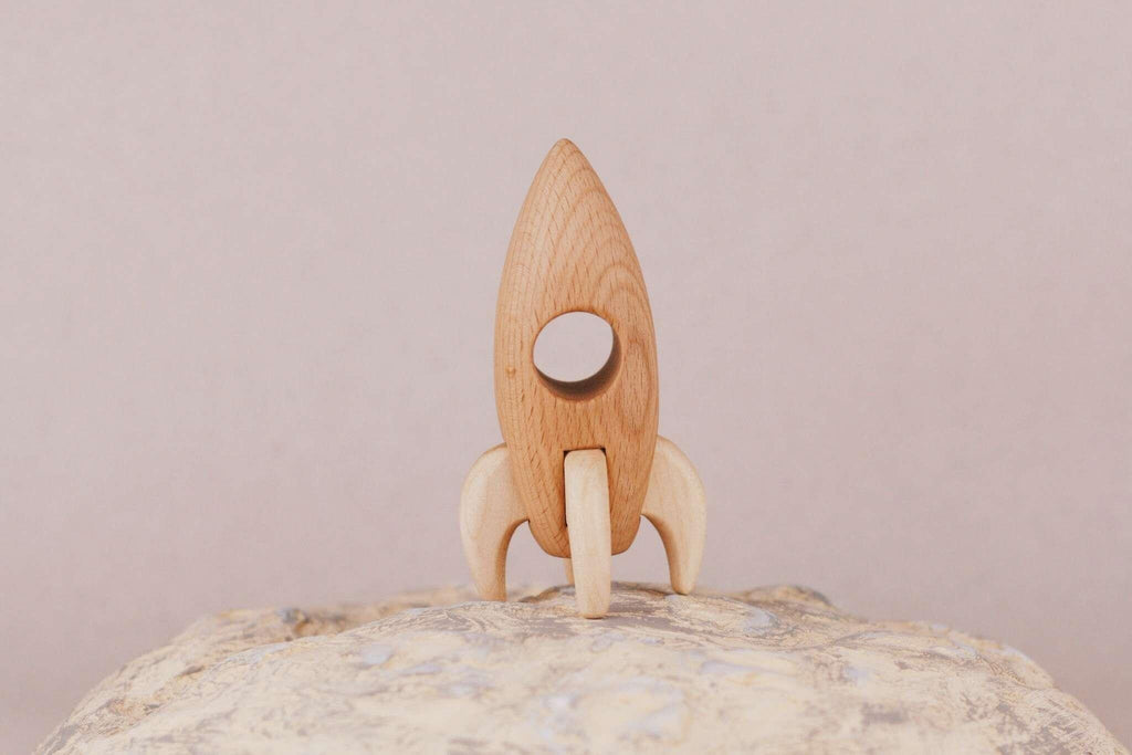 A Handmade Wooden Rocket crafted from beech wood, complete with a circular window, stands gracefully on a textured rock surface against a plain backdrop. It boasts smooth, natural wood tones accentuated by linseed oil and features simple detailing.