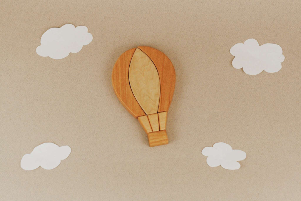 The Handmade Hot Air Balloon Puzzle, crafted from wood and finished with linseed oil, sits on a beige background. Its minimalist design is accentuated by four white paper clouds, creating a whimsical and serene atmosphere.