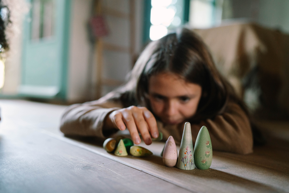 A young girl lies on the floor, focusing intently as she arranges the Grapat Play Set - Forest Tree in front of her. The background is softly blurred, creating a cozy indoor atmosphere.