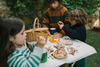Three children gather outdoors, sorting the Grapat Play Set - Mushroom Grove. A wicker basket of wooden mushrooms, bowls, and mugs sits on the table amid greenery, inspiring open-ended play and imaginative exploration.