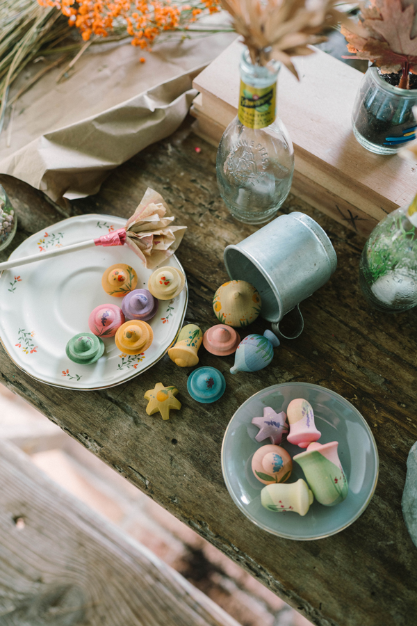 A rustic wooden table displays the Grapat Play Set - Flower Meadow with wooden mushrooms and stars on two plates, a small metal bucket, glass bottles filled with flowers, and dried botanicals in the background.