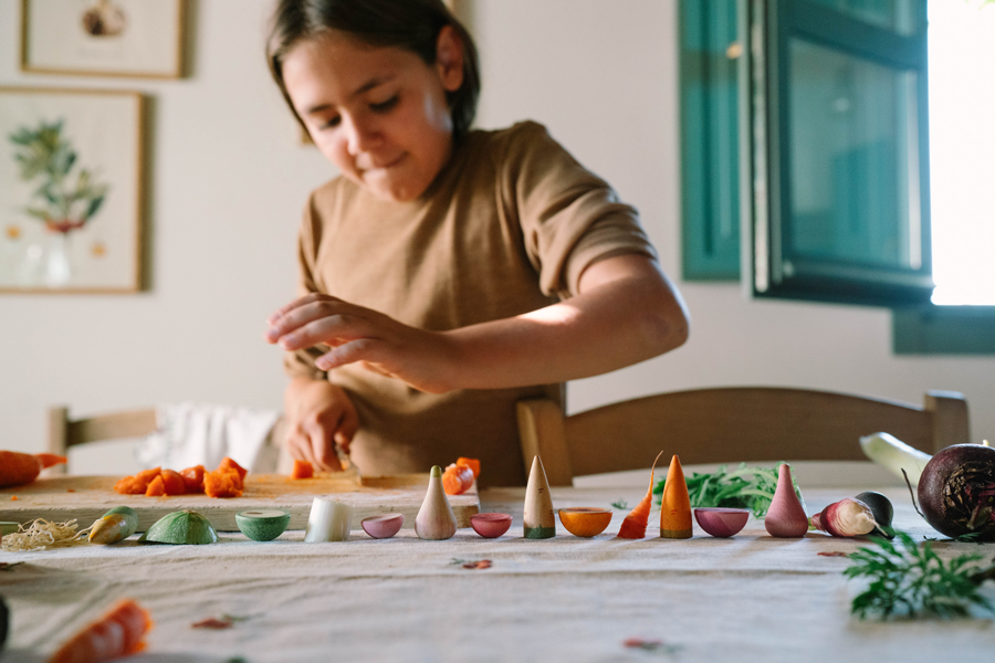 A child plays with the Grapat Play Set - Veggies, cutting wooden vegetables on a table lined with veggie pieces, a cutting board, and green herbs—perfect for creative pretend play.