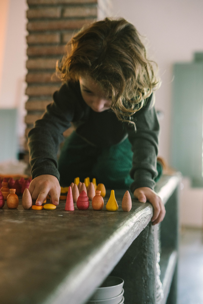 A young child with wavy hair kneels on a table, carefully arranging colorful pieces from the Grapat Mandala Mix - Warm set in neat rows. The softly lit scene has a blurred background and a warm, cozy atmosphere.