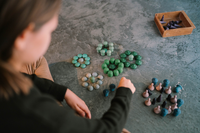 A person sits on a gray floor arranging vibrant Grapat Mandala Mix - Cold pieces and peg dolls into groups, with a wooden tray of additional sustainable wooden toys nearby. The focus is on their hand and the colorful mandalas.