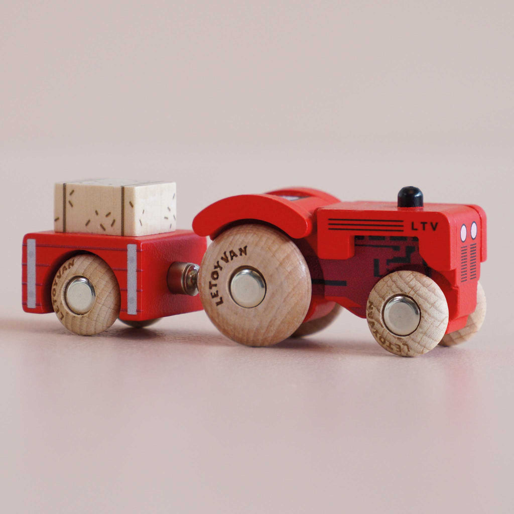 A red vintage wooden tractor labeled “No. 34 GT190 Tilly | Made in England” pulls a matching trailer carrying a small wooden block, perfect for imaginative play. Both feature natural wood wheels with visible text, set against a plain light background.