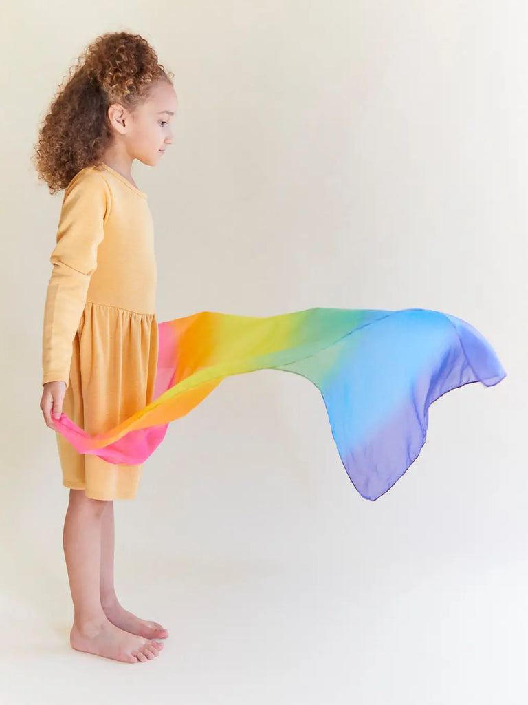 A young girl with curly hair, wearing a yellow dress, holds one end of a flowing Sarah's Silk Enchanted Playsilk - Rainbow that billows to her side against a light background.