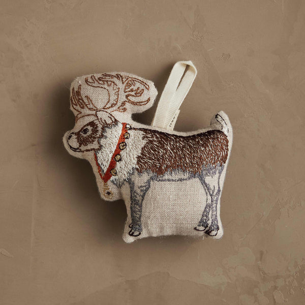 The Coral & Tusk Reindeer with Bells Ornament features embroidered details, a red collar with golden bells, and a white hanging loop, displayed on a textured beige surface.