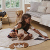 A young girl sits on a round rug with a dog face, holding a stuffed dog and reaching for a book in a cozy, sunlit bedroom featuring handcrafted decor and the Dog Cushion - Cooper.