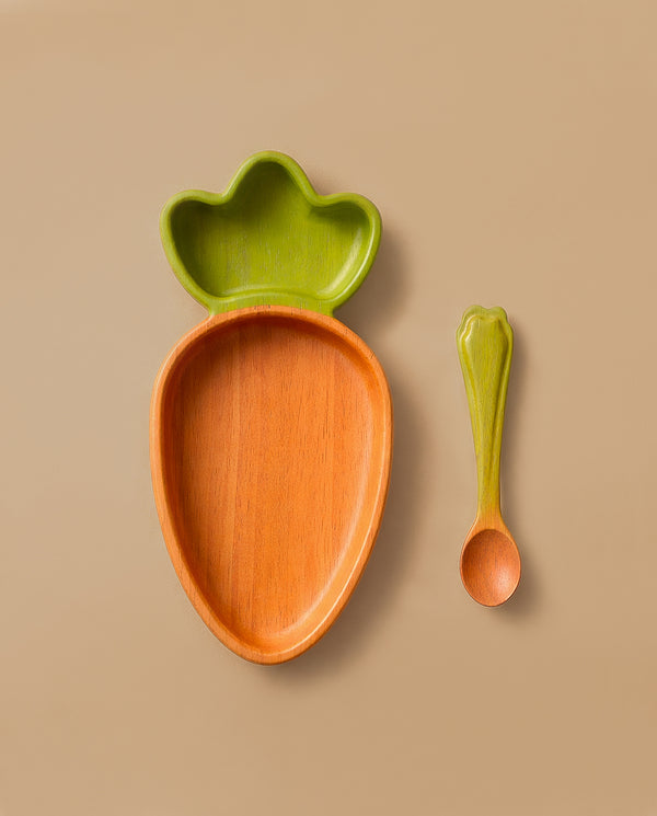 The Cathy the Carrot Plate & Spoon Set features an adorable Hevea wood plate shaped like a carrot with a green leafy top, paired with a matching green spoon—an inviting and non-toxic choice for your baby’s tableware set, shown here on a beige background.