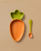 The Cathy the Carrot Plate & Spoon Set features an adorable Hevea wood plate shaped like a carrot with a green leafy top, paired with a matching green spoon—an inviting and non-toxic choice for your baby’s tableware set, shown here on a beige background.