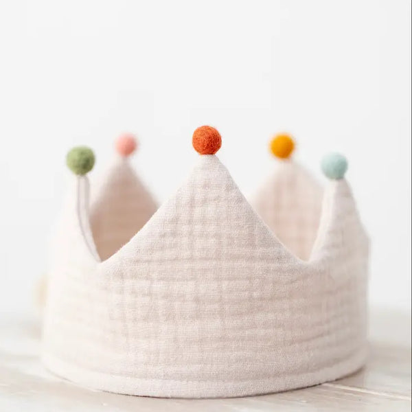 A Sand Reversible Birthday Crown with pastel-colored pom poms on each peak, set against a light, blurred background. The crown is light grey, handmade from organic cotton, evoking a playful.