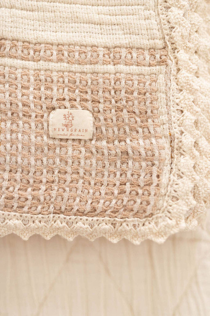 Close-up of the Organic Cotton Baby Blanket - Farm, featuring textured beige woven fabric, a crochet trim, and a centered label with "NEVER AGAIN" and a decorative logo above.