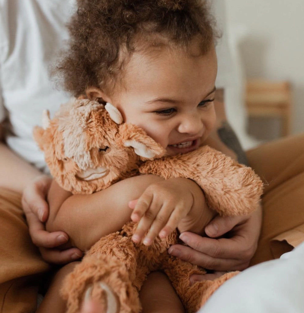 A toddler with curly hair smiling and hugging a Slumberkins Yak Kin while sitting on an adult's lap, conveying a sense of comfort and joy.