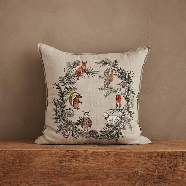 The Coral & Tusk Tree Trimmers Wreath Pillow, in beige with embroidered woodland animals like a fox, bear, owl, rabbit, squirrel, and goat among pine branches and cones, adds festive charm to your seasonal decor.