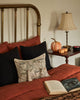 A cozy bed with rust-colored bedding and black pillows features the Coral & Tusk Graveyard Pocket Pillow (ships in one week) near a brass headboard. By the bed are a lamp, stacked books, decorative pumpkins, and an open book.