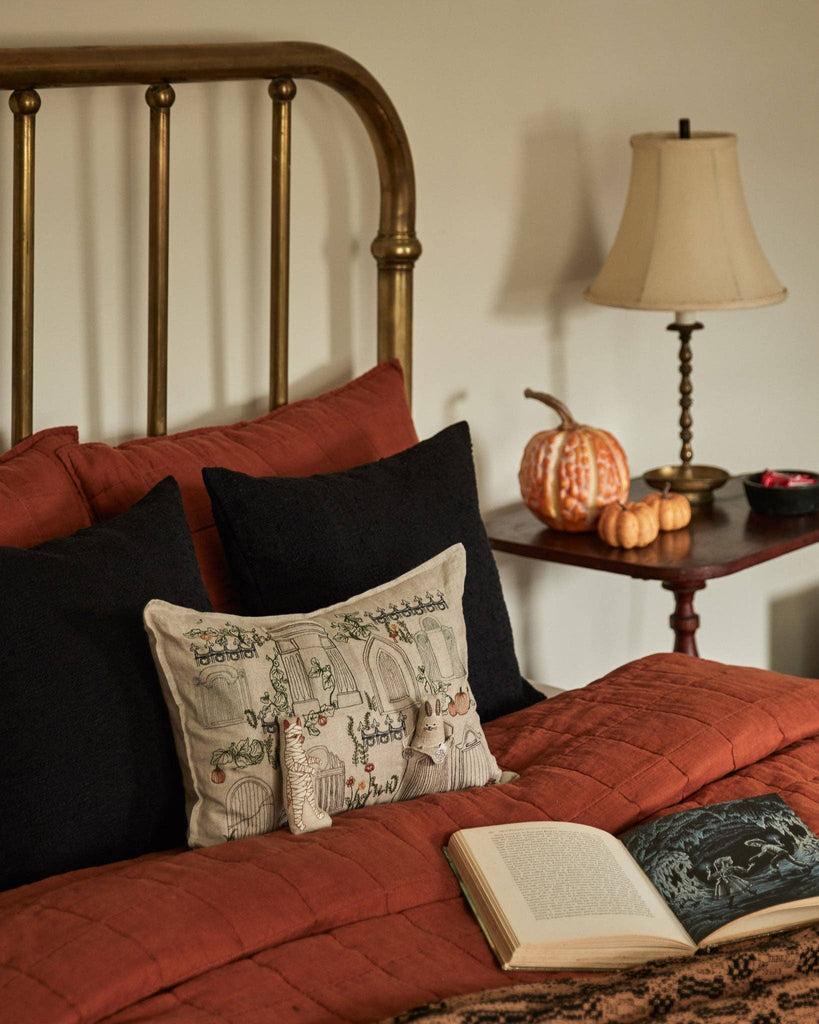 A cozy bed with rust-colored bedding and black pillows features the Coral & Tusk Graveyard Pocket Pillow (ships in one week) near a brass headboard. By the bed are a lamp, stacked books, decorative pumpkins, and an open book.