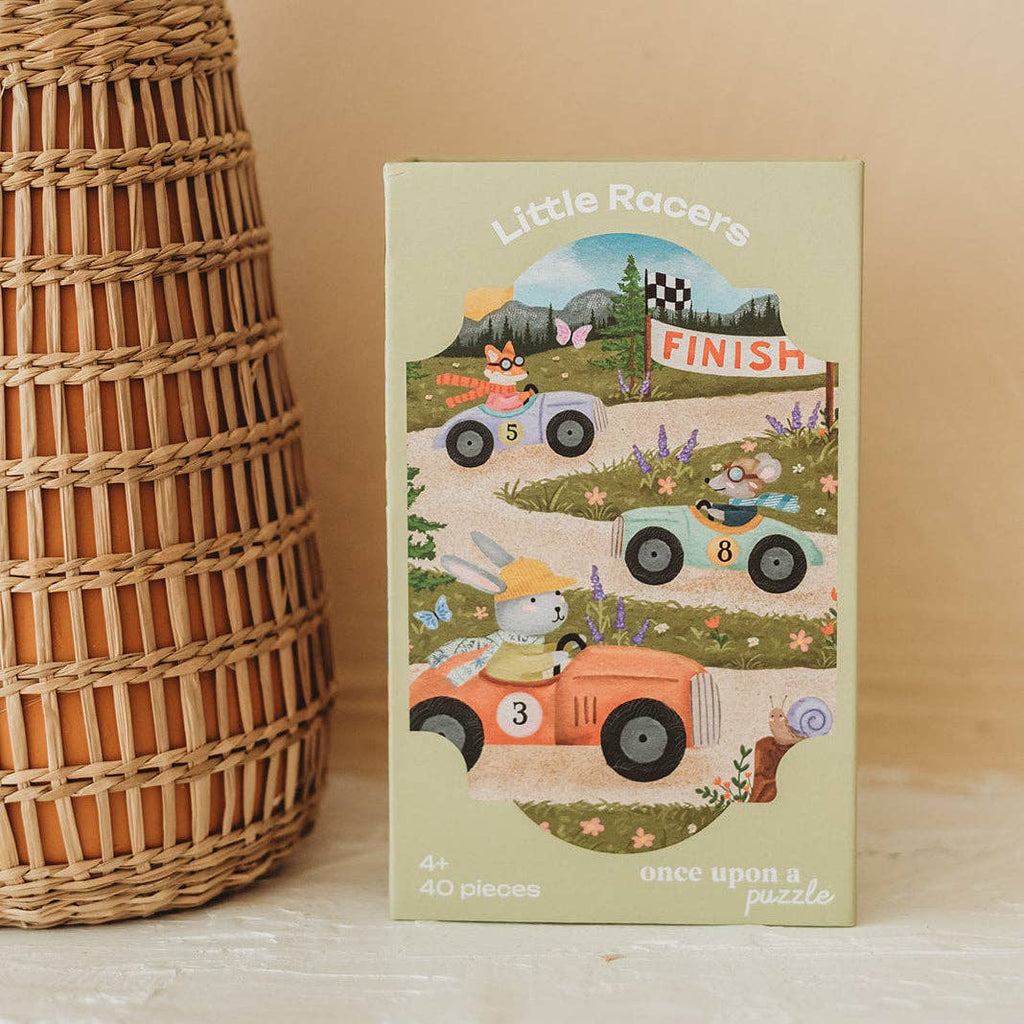 The Little Racers 40 Piece Mini puzzle box features illustrated woodland animals racing cars toward a finish line. Designed for children ages 4 and up, the box is labeled with “40 pieces” and “once upon a puzzle.” Set beside a woven basket, it invites young puzzlers to join in the fun.