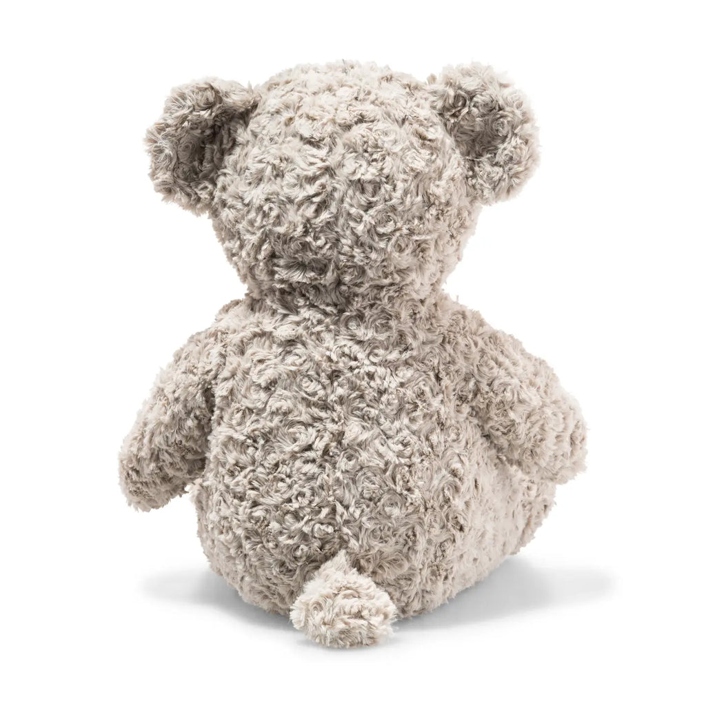 A rear view of the fluffy, 19-inch gray Steiff Honey Teddy Bear sitting upright. As part of the Soft Cuddly Friends Collection, it features curly fur, round ears, and a small tail. The white background accentuates the toy's soft texture and cuddly charm.