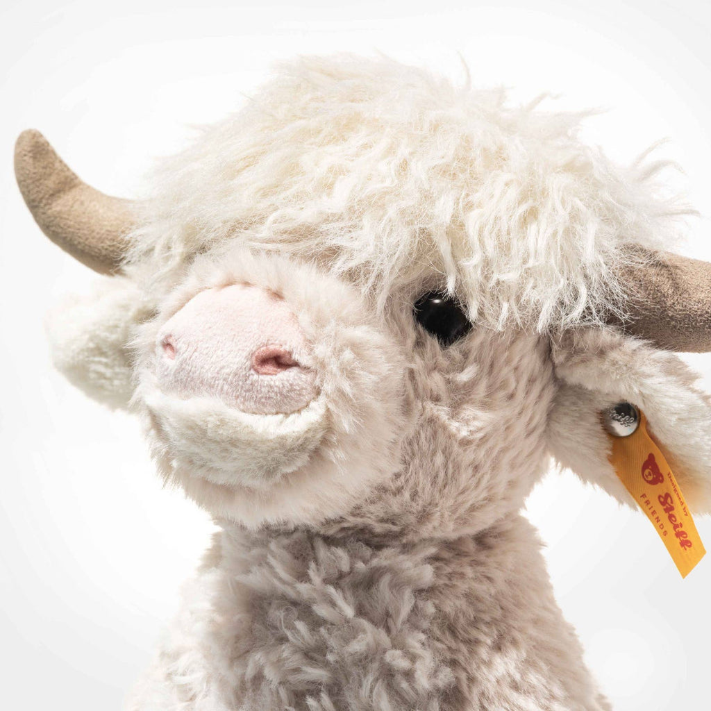 The Steiff Harper Highland Cow is a fluffy, cream-colored plush with big beige horns, a pink nose, and the signature yellow Steiff ear tag—ideal as a cozy nursery toy—set against a plain white background.
