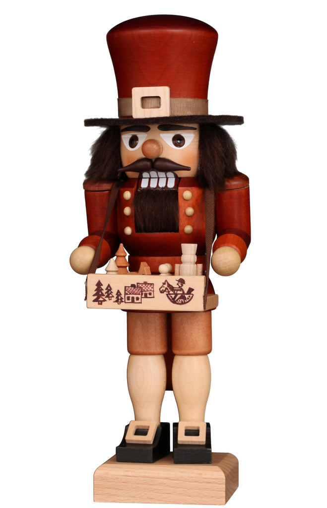 The Christian Ulbricht Collectible Wooden Nutcracker - Toy Trader features a tall red hat, brown beard and mustache, and holds a box adorned with forest and village scenes containing small wooden items.