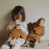 A young child sits against a beige wall, hugging a large Senger Naturwelt Cuddly Animal - Rust Goose. Another similar organic cotton goose rests beside them as the child looks away, partially hiding their face.
