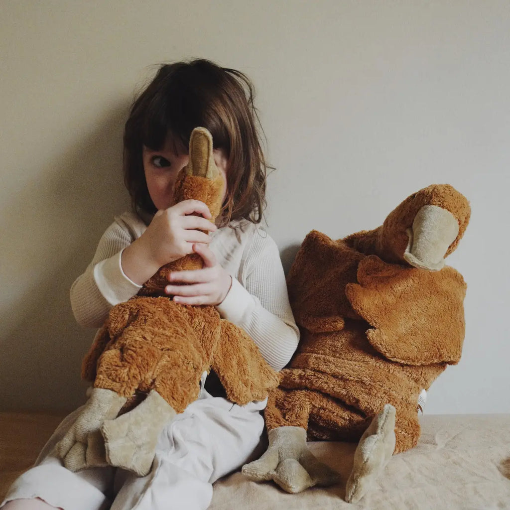 A young child sits against a beige wall, hugging a large Senger Naturwelt Cuddly Animal - Rust Goose. Another similar organic cotton goose rests beside them as the child looks away, partially hiding their face.