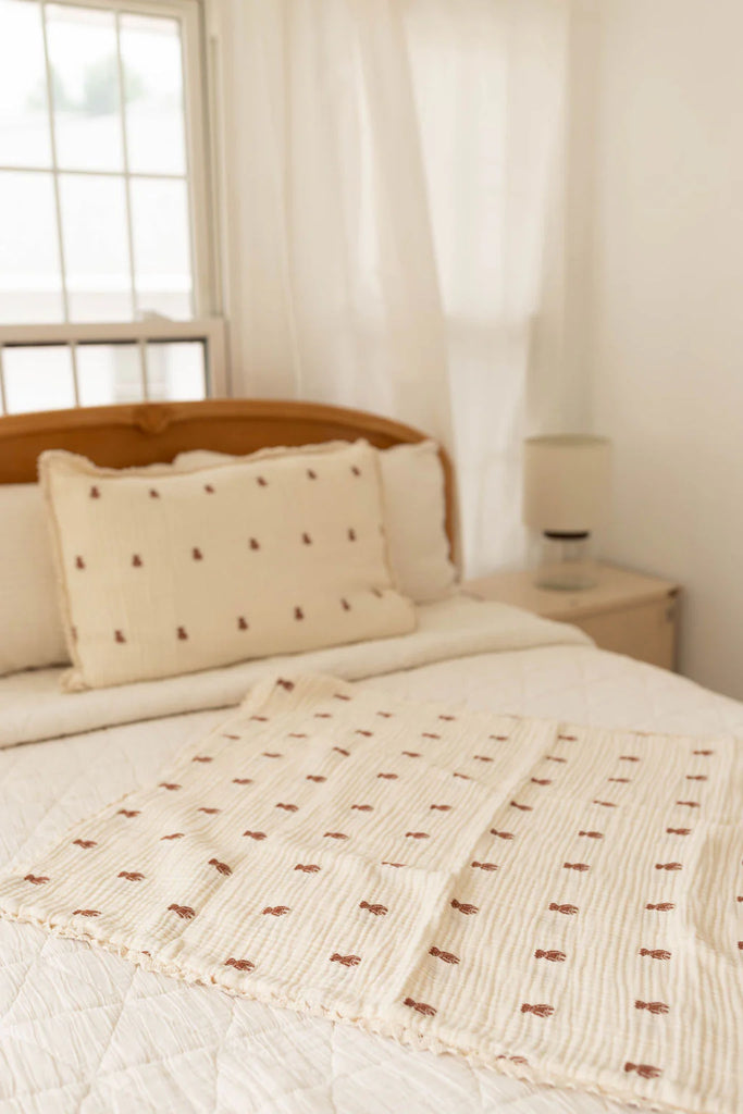 A neatly made bed with cream bedding featuring a brown dot pattern, a matching pillow, and wooden headboard is topped with the Organic Cotton Layered Gauze Baby Blanket - Teddy. A bedside table with lamp sits by a window in sheer white curtains.
