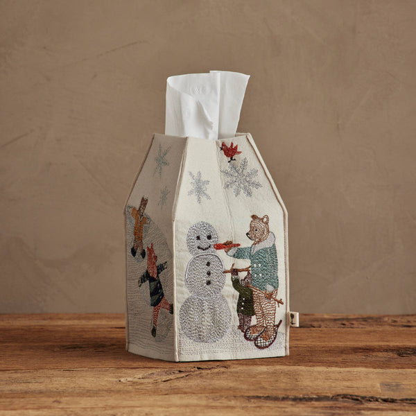 The Coral & Tusk Snow Day Tissue Box Cover, featuring embroidered animals building a snowman, adds charming snow day decor and sits on a wooden surface with a neutral beige background.