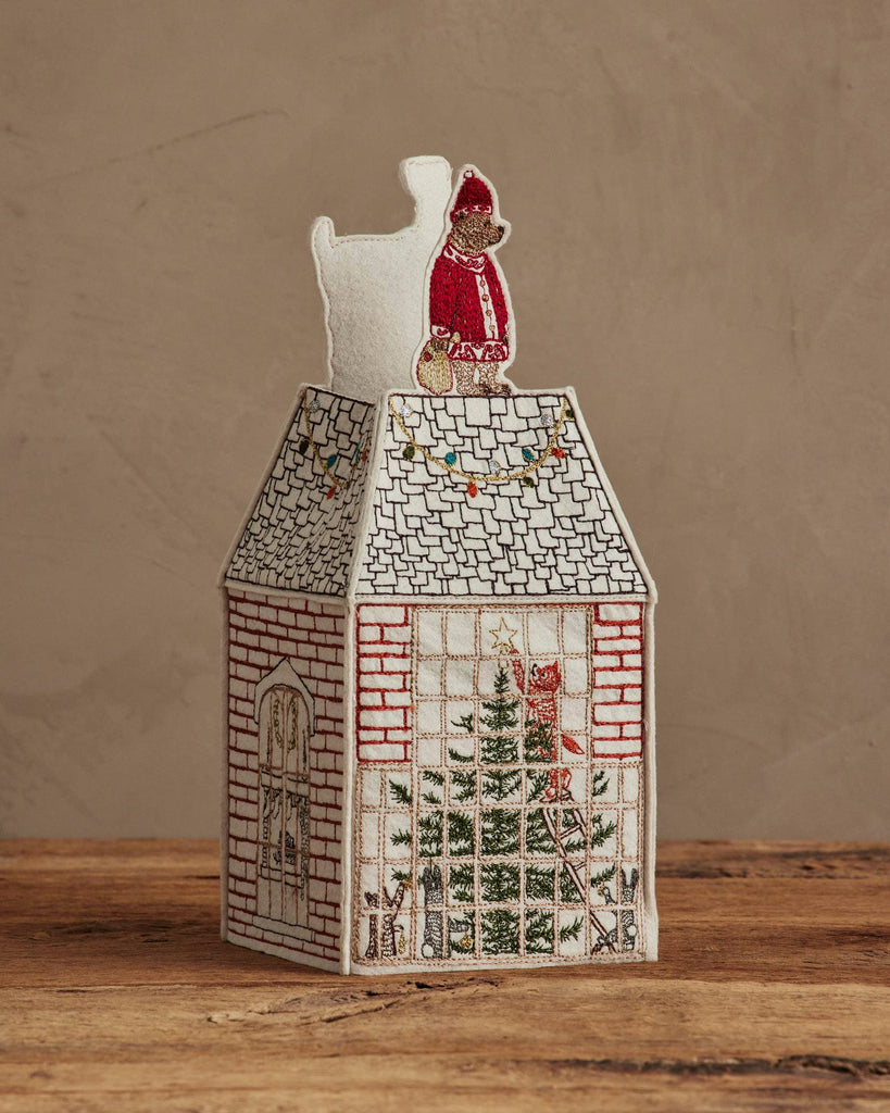 The Coral & Tusk Home for the Holidays Tissue Box Cover is a house-shaped, embroidered cover with brick walls, windows, a Christmas tree inside, and a red-clad figure on the roof, displayed on wood against a neutral background.