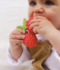 Wearing a white shirt and brown overalls, a baby chews on Valery the Raspberry Teething Toy—a red, raspberry-shaped natural rubber teether with green leaves. The eco-friendly toy captures the baby’s curiosity as they gaze upward.